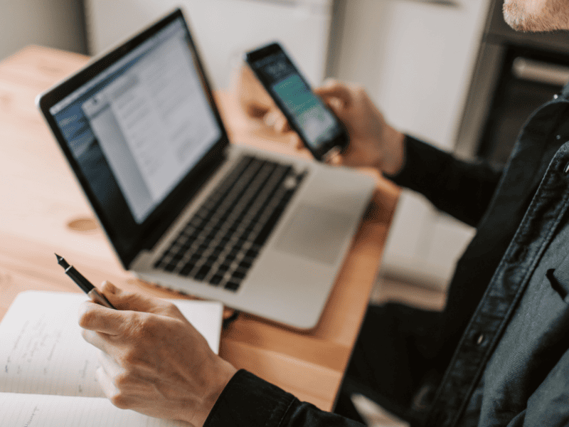 Pessoa usando um notebook enquanto segura um smartphone e faz anotações em um caderno sobre a mesa.