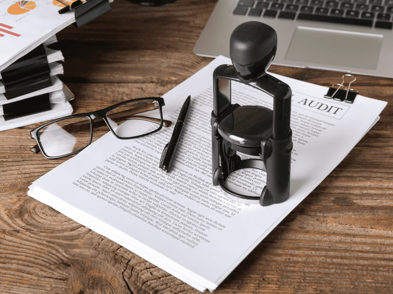 Carimbo de cartório sobre documentos impressos, ao lado de óculos, caneta e um notebook sobre a mesa.