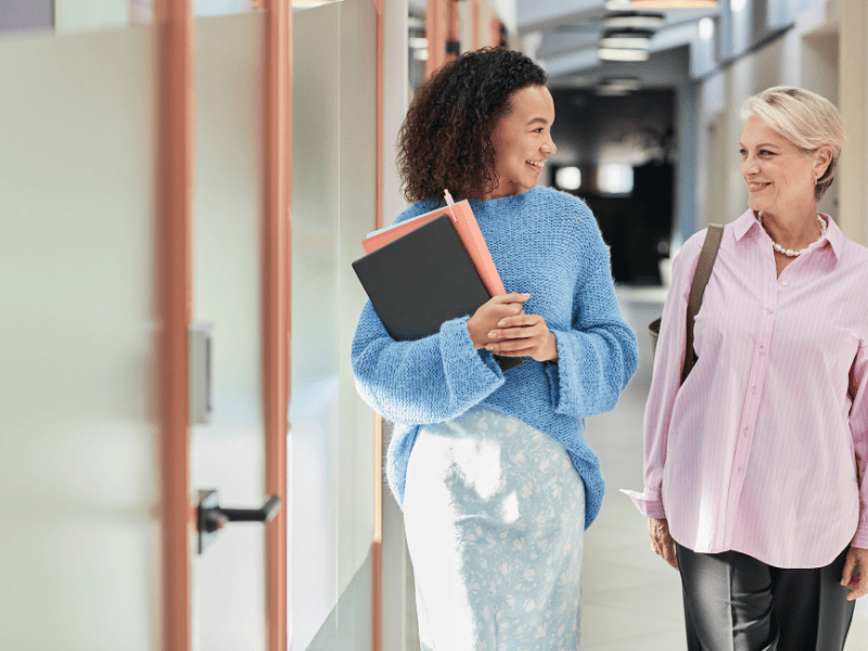 Duas mulheres caminhando por um corredor enquanto conversam; uma delas carrega cadernos.