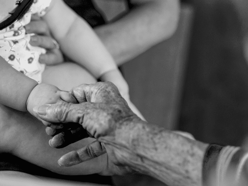 Mão de uma pessoa idosa segurando a mão de um bebê em uma imagem em preto e branco.