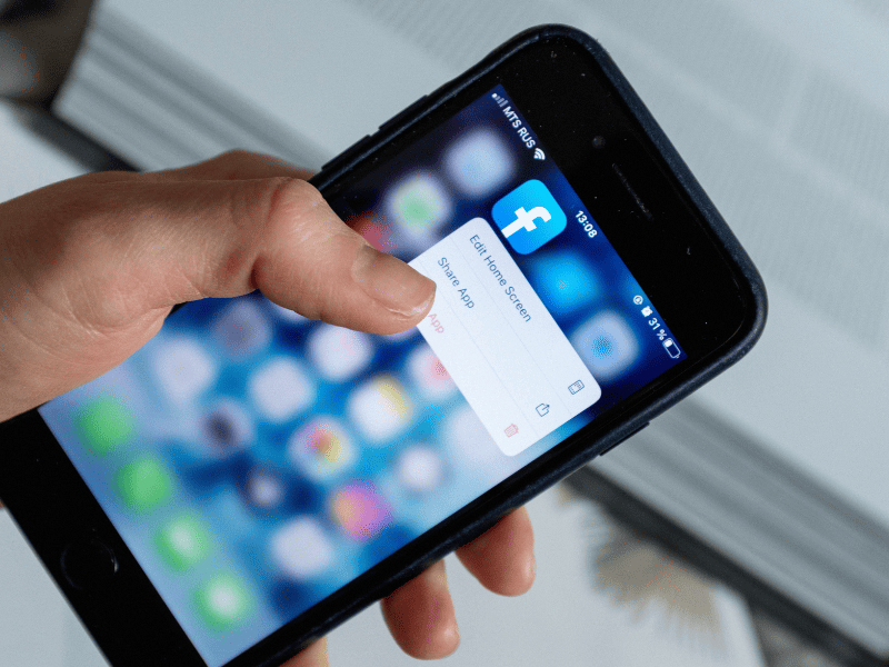 Pessoa segurando celular com o ícone do Facebook pressionado
