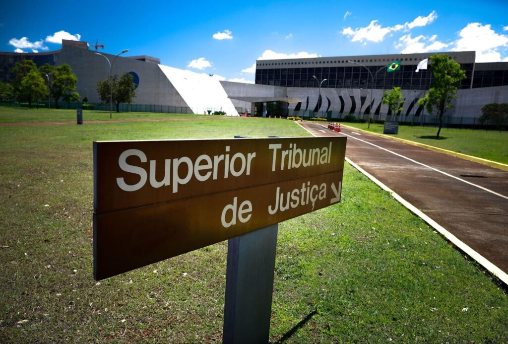 Placa do Superior Tribunal de Justiça em área externa, com o prédio do tribunal ao fundo.