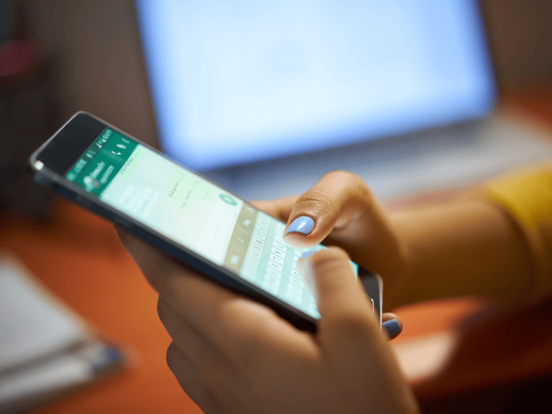 Pessoa digitando mensagens no WhatsApp em um smartphone, com um notebook ao fundo.