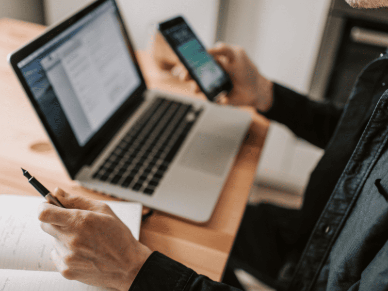 Pessoa segurando um smartphone com o WhatsApp aberto enquanto usa um notebook e faz anotações em um caderno.