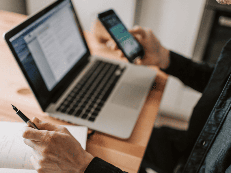 Pessoa segurando um smartphone enquanto usa um notebook e faz anotações em um caderno.