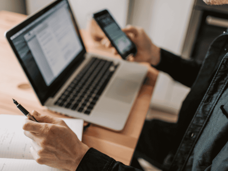 Pessoa segurando um celular enquanto utiliza um notebook sobre a mesa, com documentos abertos na tela e em papel.
