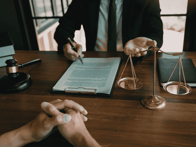 Pessoa sentada à mesa com documentos impressos, caneta na mão, balança da justiça e martelo de juiz sobre a mesa.