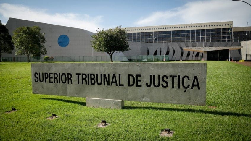 Placa com a inscrição “Superior Tribunal de Justiça” em frente ao prédio do tribunal, com área verde e fachada institucional ao fundo.