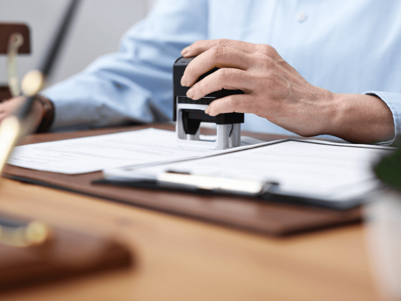 Pessoa pressionando um carimbo sobre documentos impressos em uma mesa, com papéis e prancheta ao fundo.