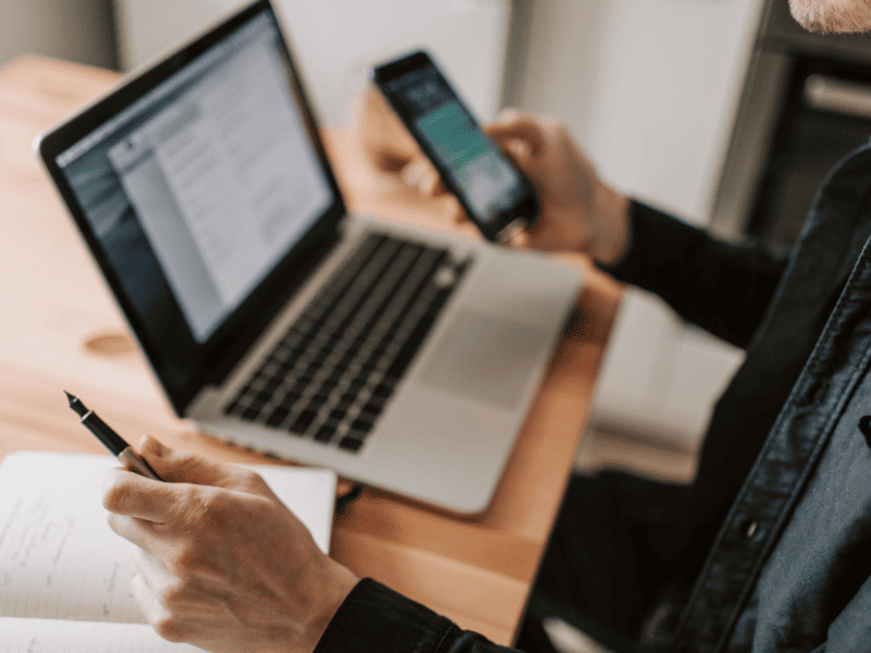 Pessoa segurando um smartphone enquanto usa um notebook e faz anotações em um caderno.