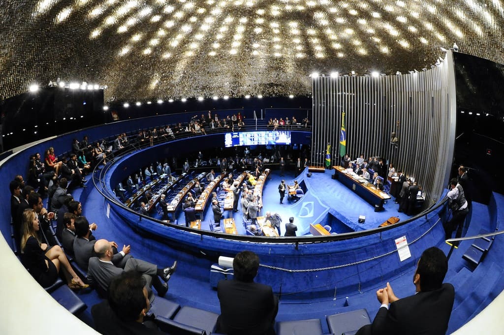 Plenário do Senado Federal com parlamentares reunidos em sessão, com público nas galerias e bandeira do Brasil ao fundo.
