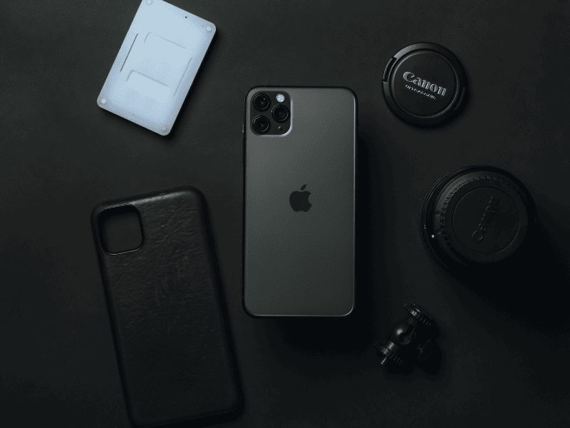 Smartphone iPhone sobre mesa escura ao lado de capa, acessórios e lentes de câmera Canon
