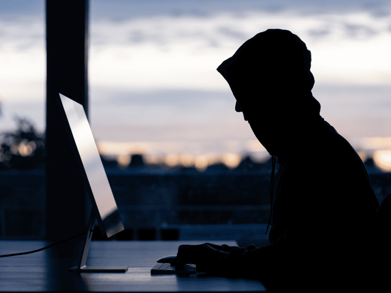 Silhueta de pessoa com capuz usando computador em frente a uma janela, com cidade ao fundo ao entardecer