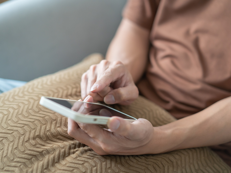 Pessoa sentada segurando um smartphone, tocando a tela com o dedo enquanto usa o aparelho sobre uma almofada