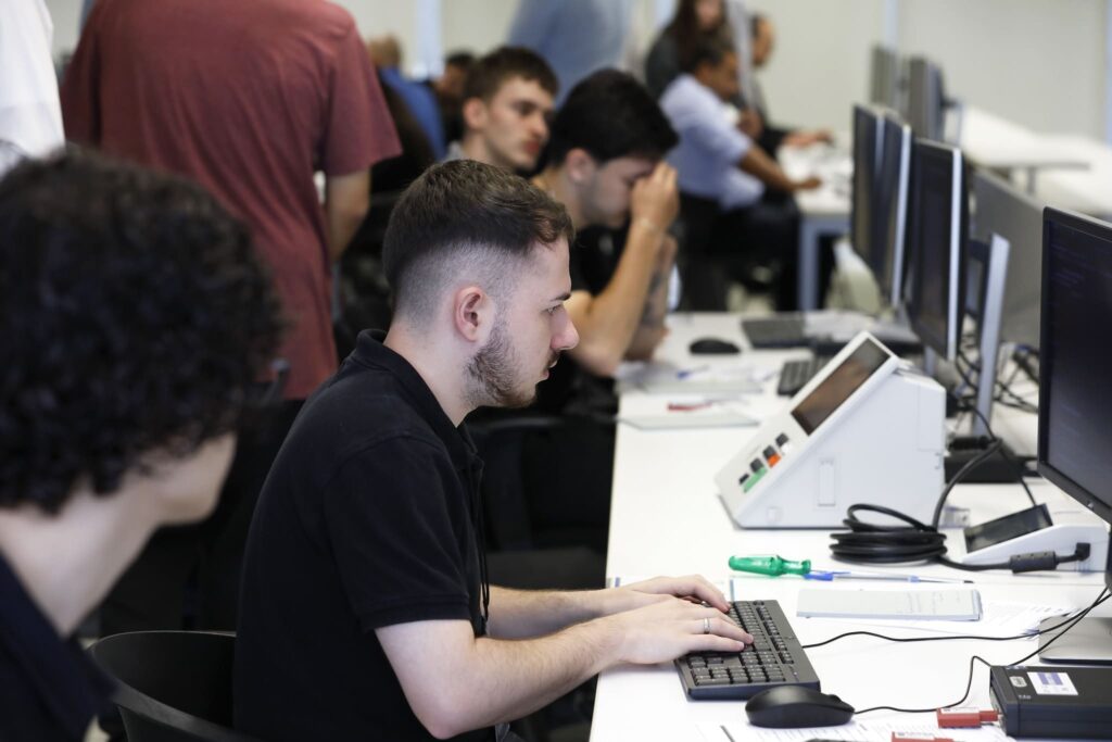 Equipe trabalhando em computadores e equipamentos eletrônicos em ambiente de testes eleitorais.