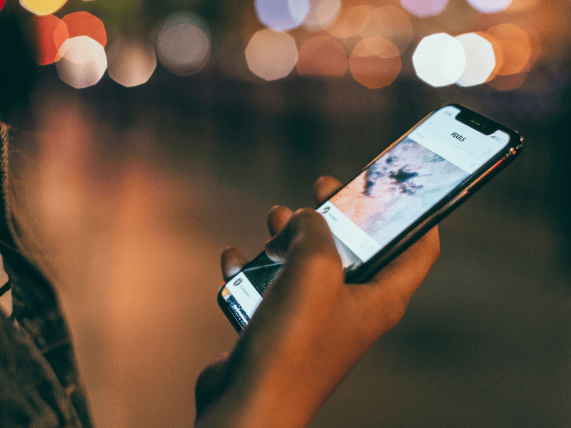 Pessoa segurando smartphone com rede social aberta na tela, com luzes desfocadas ao fundo durante a noite.