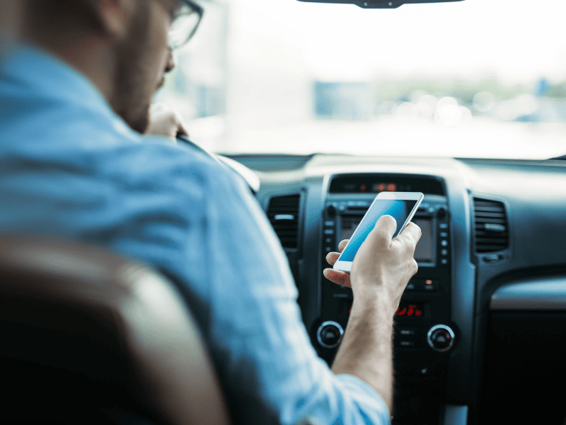 Homem dentro de um carro, sentado no banco do motorista, segurando um smartphone com a tela iluminada, enquanto o painel do veículo aparece ao fundo.