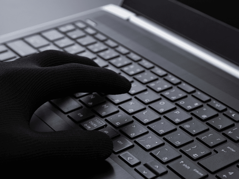 Mão com luva preta digitando em teclado de notebook em close, com iluminação escura.