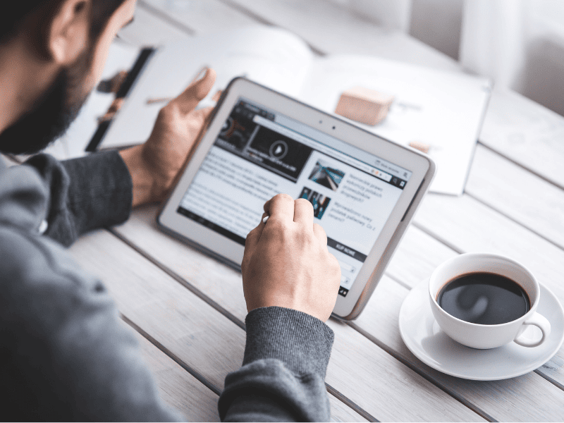 Pessoa utilizando um tablet para navegar em um site de notícias enquanto toma café em uma mesa.