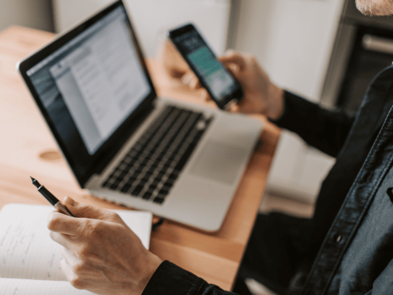 Pessoa segurando um smartphone enquanto utiliza um laptop e faz anotações em um caderno sobre a mesa.