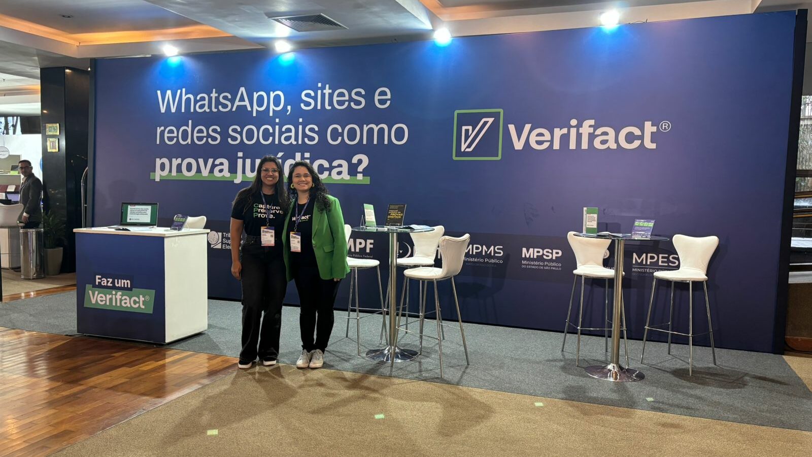 Duas integrantes da equipe Verifact posam no estande da marca no IBCCRIM 2026, com o painel "WhatsApp, sites e redes sociais como prova jurídica?" e o totem "Faz um Verifact" ao fundo